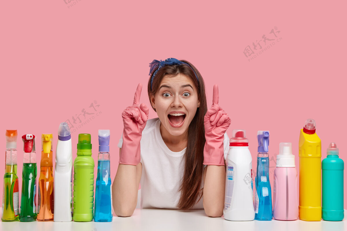 清洁剂乐观的年轻女佣用食指向上指 戴着头带和橡胶手套 在天花板上展示一些令人敬畏的东西瓶子T恤乐观