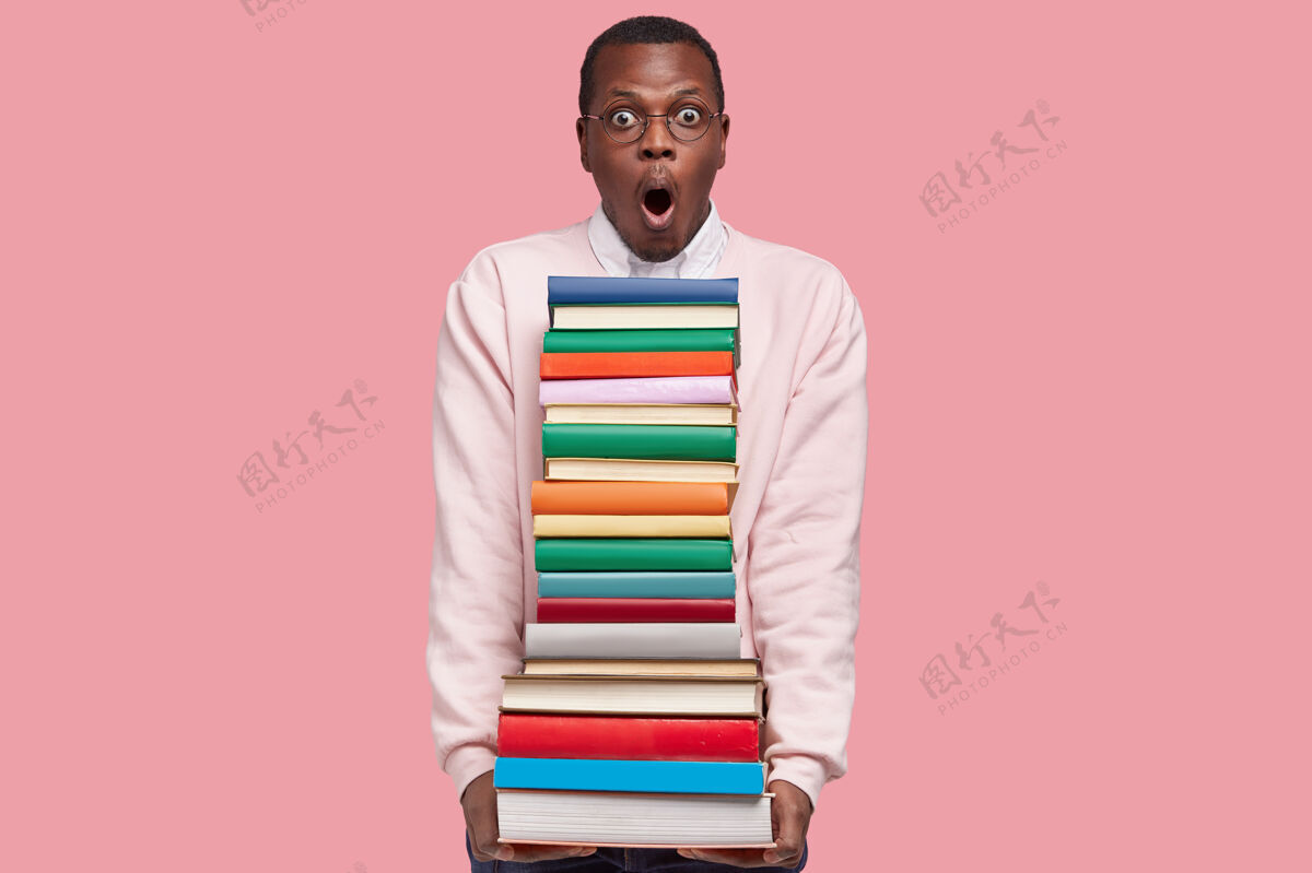 堆照片中穿着白色休闲套头衫的黑人年轻人提着一堆书 戴着圆眼镜 站在粉红色的墙上眼镜可怕教科书