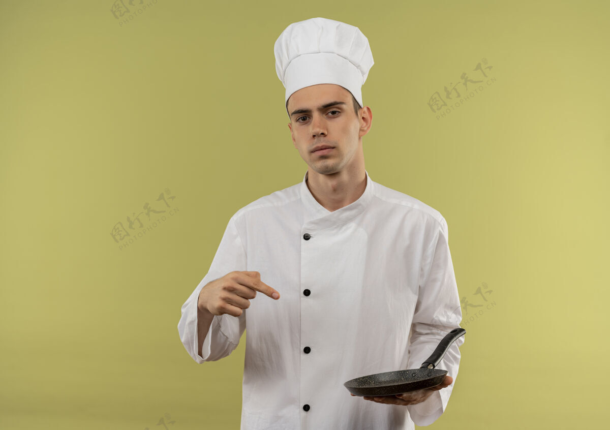 厨师身着厨师制服的自信年轻男厨师将手指指向他手上的煎锅 放在隔离的绿色墙壁上厨师男性平底锅
