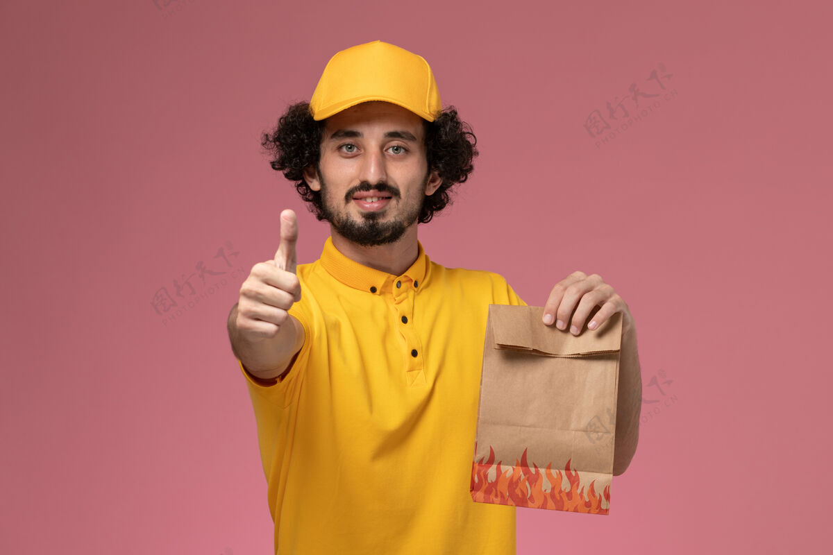 工人正面图身穿黄色制服的男快递员拿着纸质食品包在浅粉色的墙上摆姿势持有男性快递
