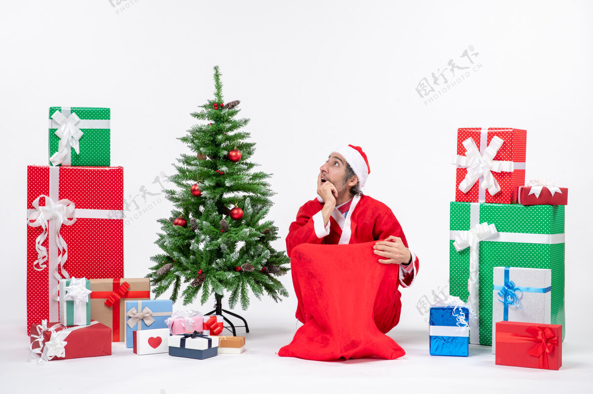 微笑困惑的年轻人打扮成圣诞老人 拿着礼物和装饰过的圣诞树坐在地上的白色背景上圣诞男人圣诞老人
