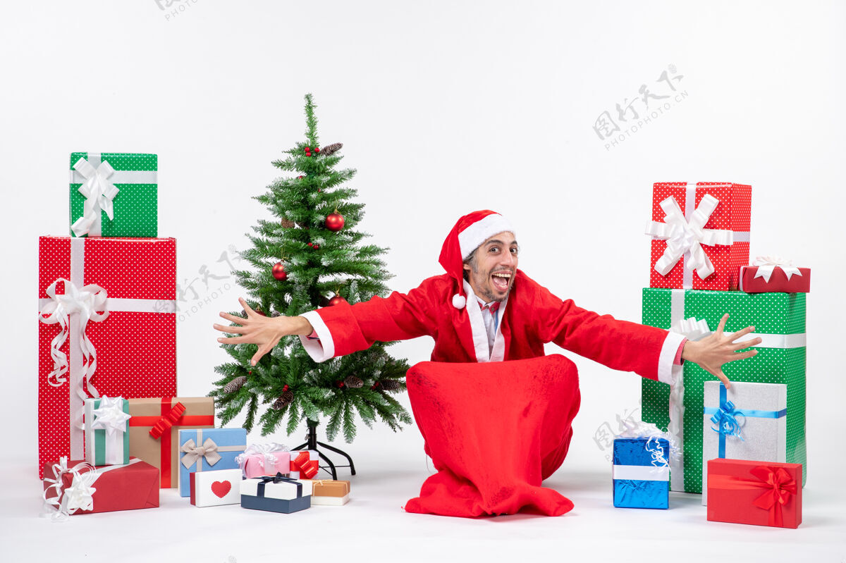 礼物滑稽快乐的年轻人打扮成圣诞老人 拿着礼物和装饰好的圣诞树坐在白色背景的地上圣诞树人年轻人