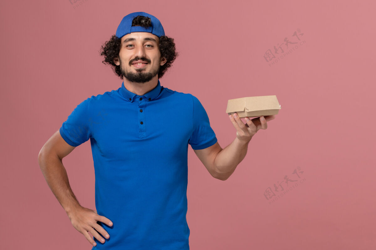 手持正面图身穿蓝色制服和披风的男性快递员拿着粉色墙上的小快递食品包粉色男信使人
