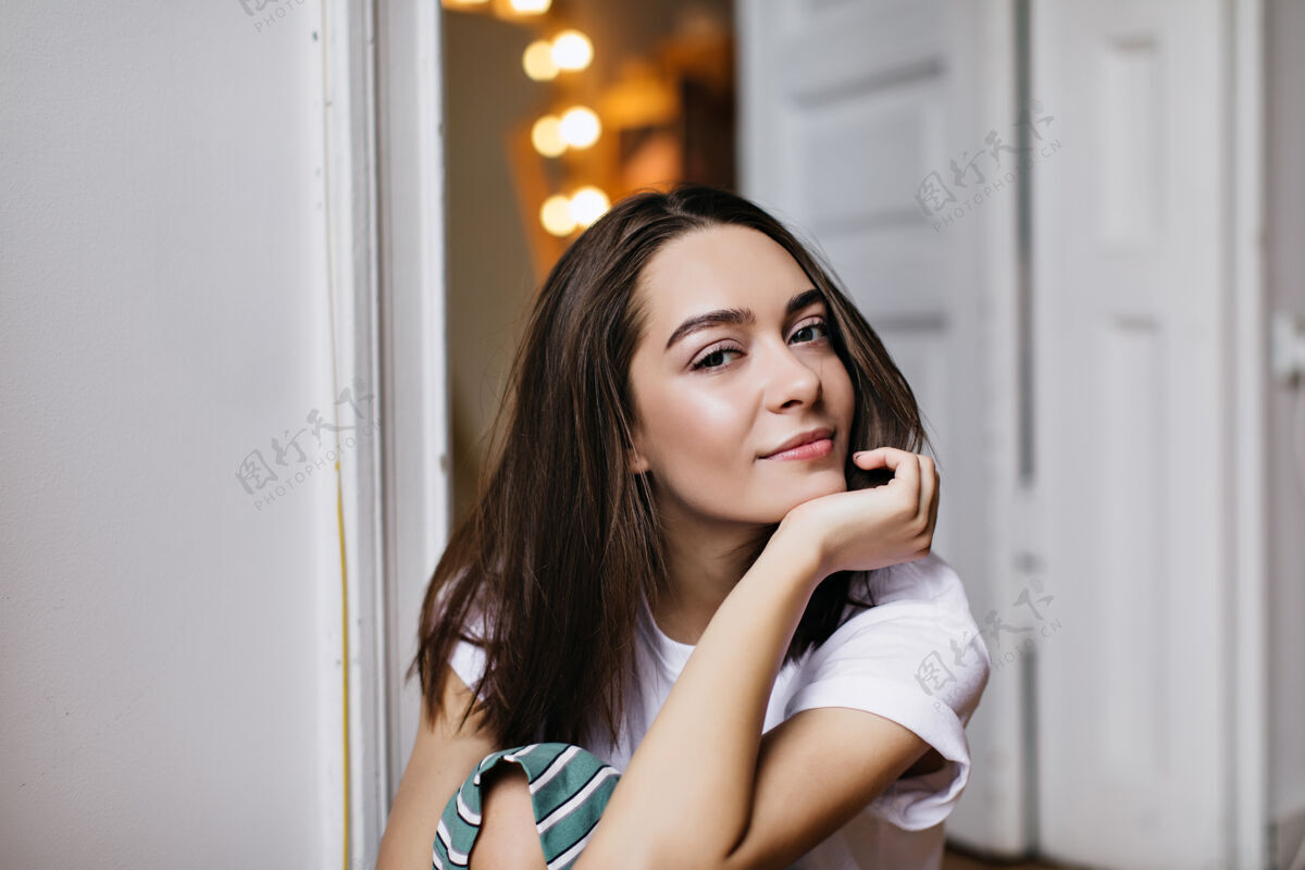 棕色头发可爱的深棕色头发的女孩在家里的白色门前摆姿势梦幻的欧洲女人抚摸着她的脸看的室内镜头休息放松美丽