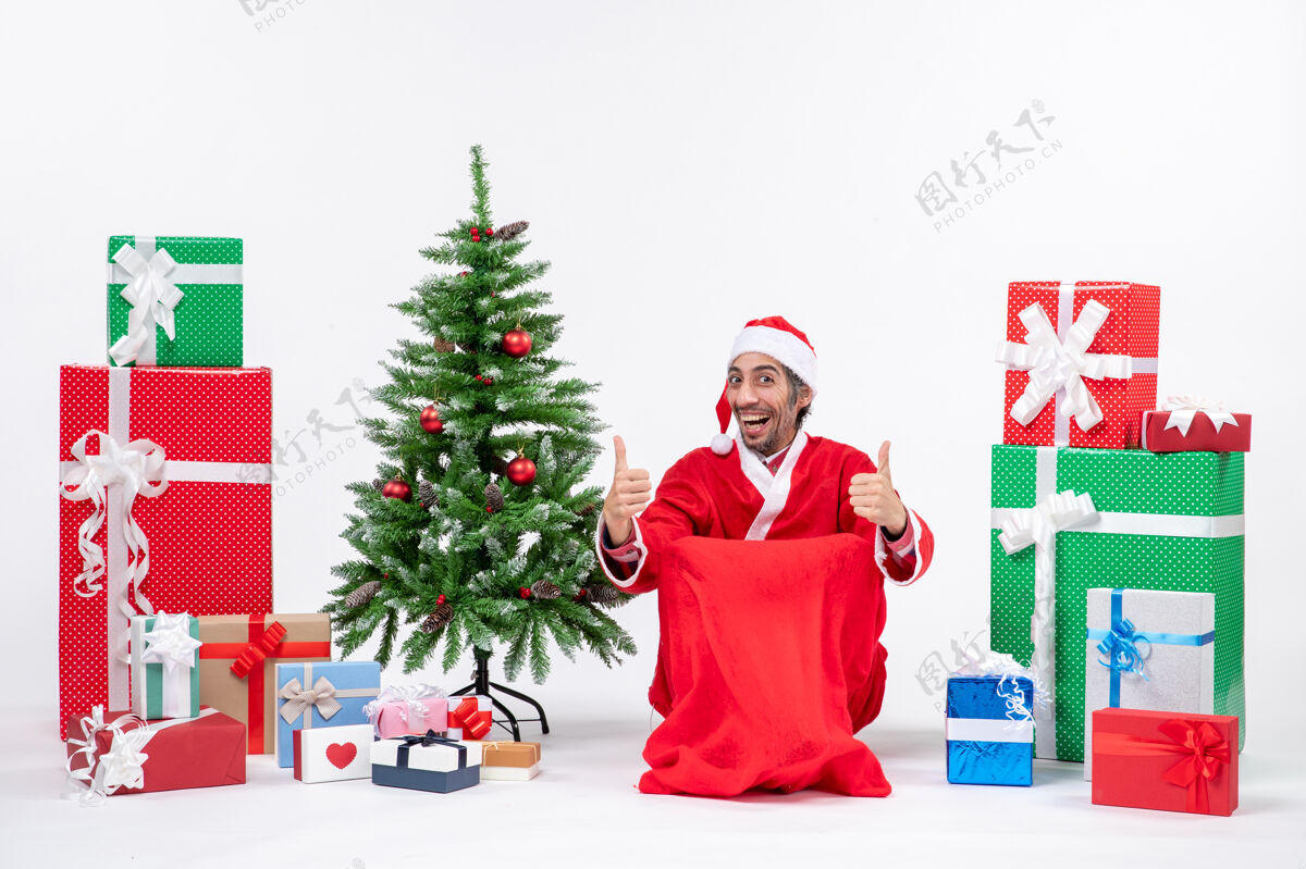 圣诞树快乐的年轻人打扮成圣诞老人与礼物和装饰圣诞树作出完美的姿态在白色背景圣诞老人帽子圣诞老人