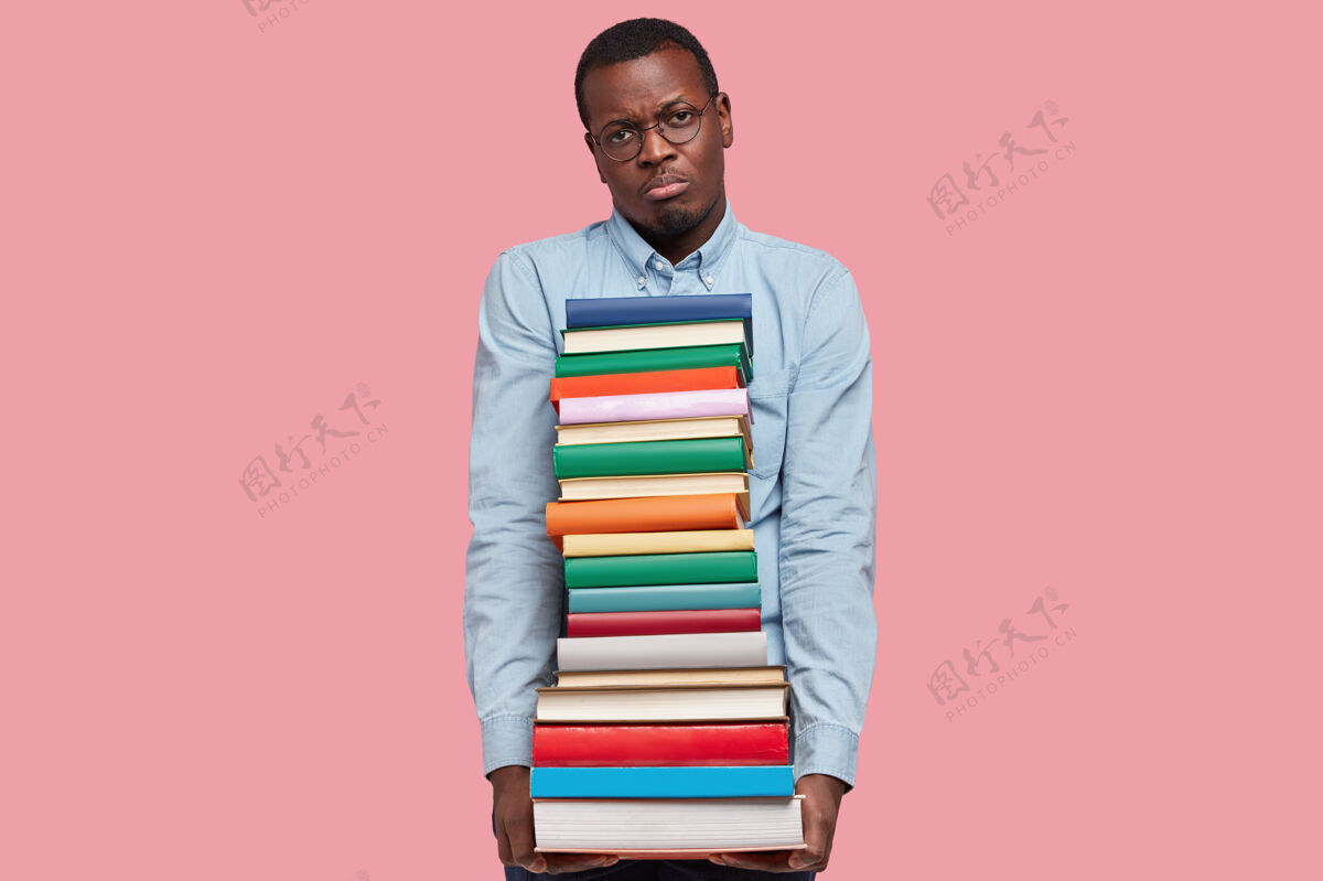 考试不高兴的黑皮肤商人的照片拿着一堆科学书籍 有着不高兴的面部表情 穿着正式的衬衫和眼镜 隔着粉红色的工作室墙冷漠暗黑皮肤个性