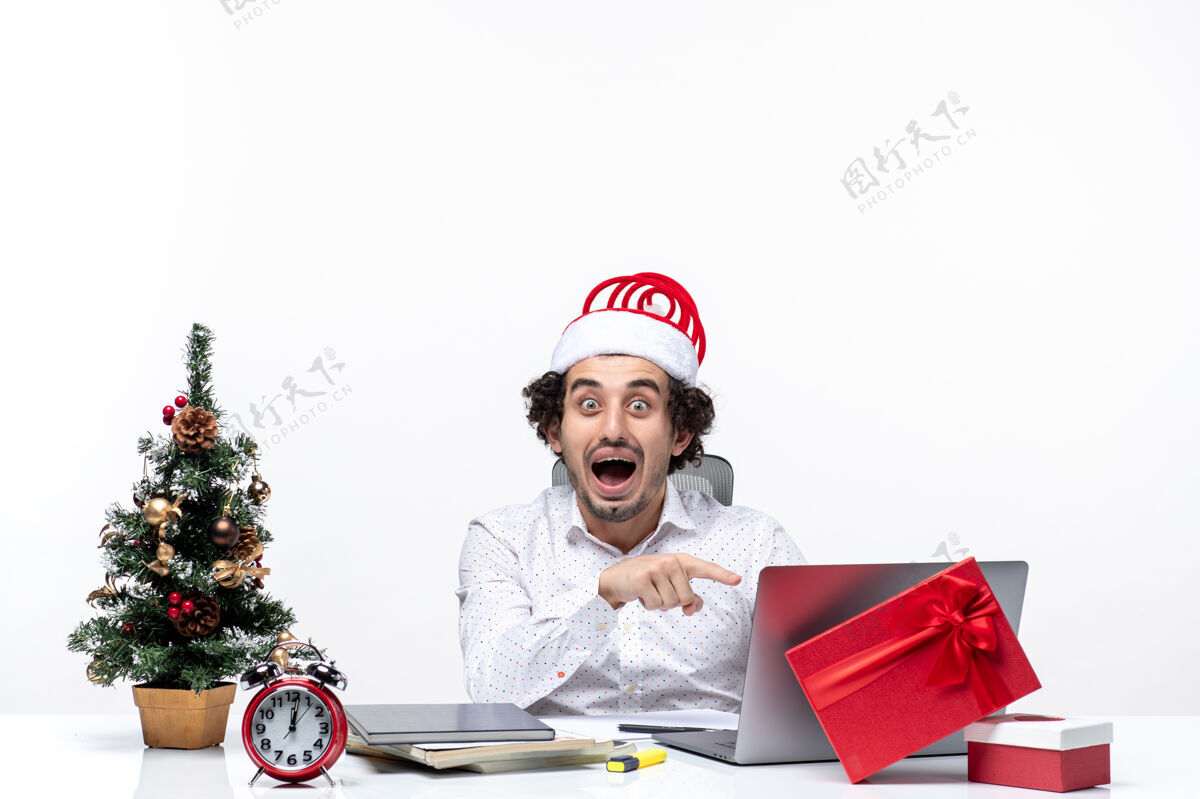 商人快乐积极的年轻商人 戴着有趣的圣诞老人帽子 在白色背景下的办公室里的笔记本电脑上展示着一些东西帽子圣诞老人圣诞老人