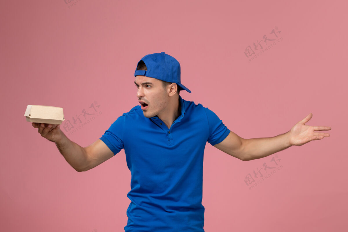 肖像正面图：身穿蓝色制服披肩的年轻男性快递员手拿着小快递食品包站在粉红色的墙上人男性持有