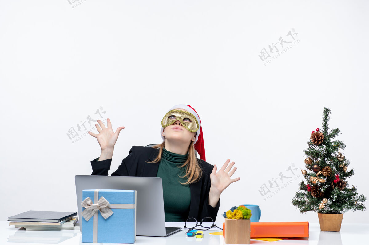 圣诞老人圣诞气氛与年轻女子戴着圣诞老人的帽子 戴着面具坐在一张桌子上看上面的白色背景桌子笔记本电脑圣诞节