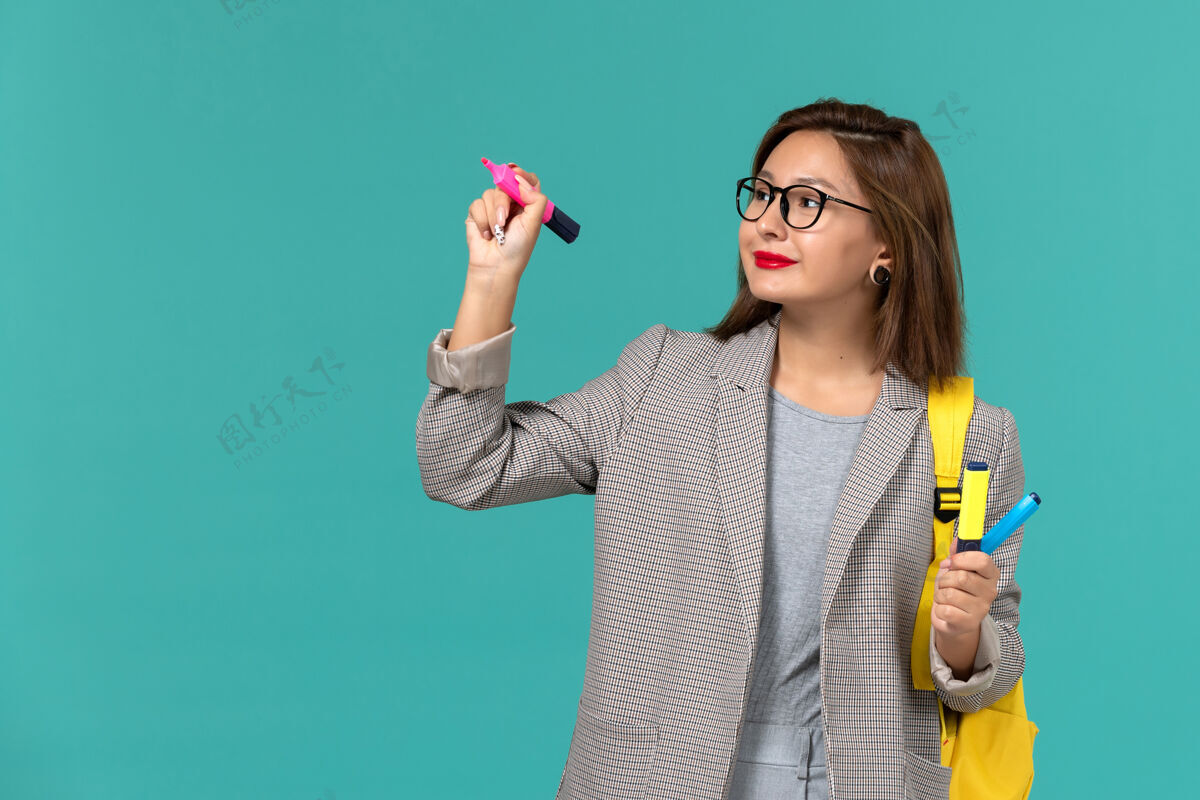 Female身穿灰色夹克 背着黄色背包 拿着毛毡笔的女学生在浅蓝色墙上的正面照片感觉人漂亮