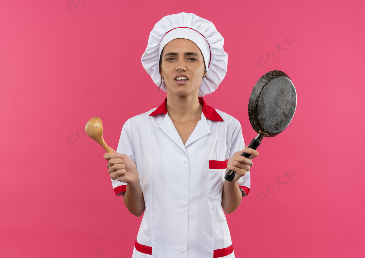 年轻困惑的年轻女厨师穿着厨师制服 拿着煎锅和勺子 站在粉红色的墙上 还有复印空间制服困惑煎炸