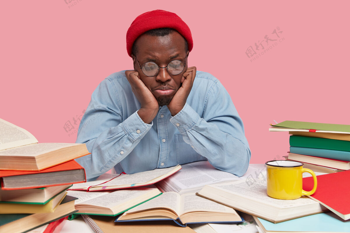 手册心烦意乱的黑人年轻人注意力不集中 长时间看书感到沮丧和悲伤 戴着圆眼镜 戴着红帽子和衬衫个人男性坐着