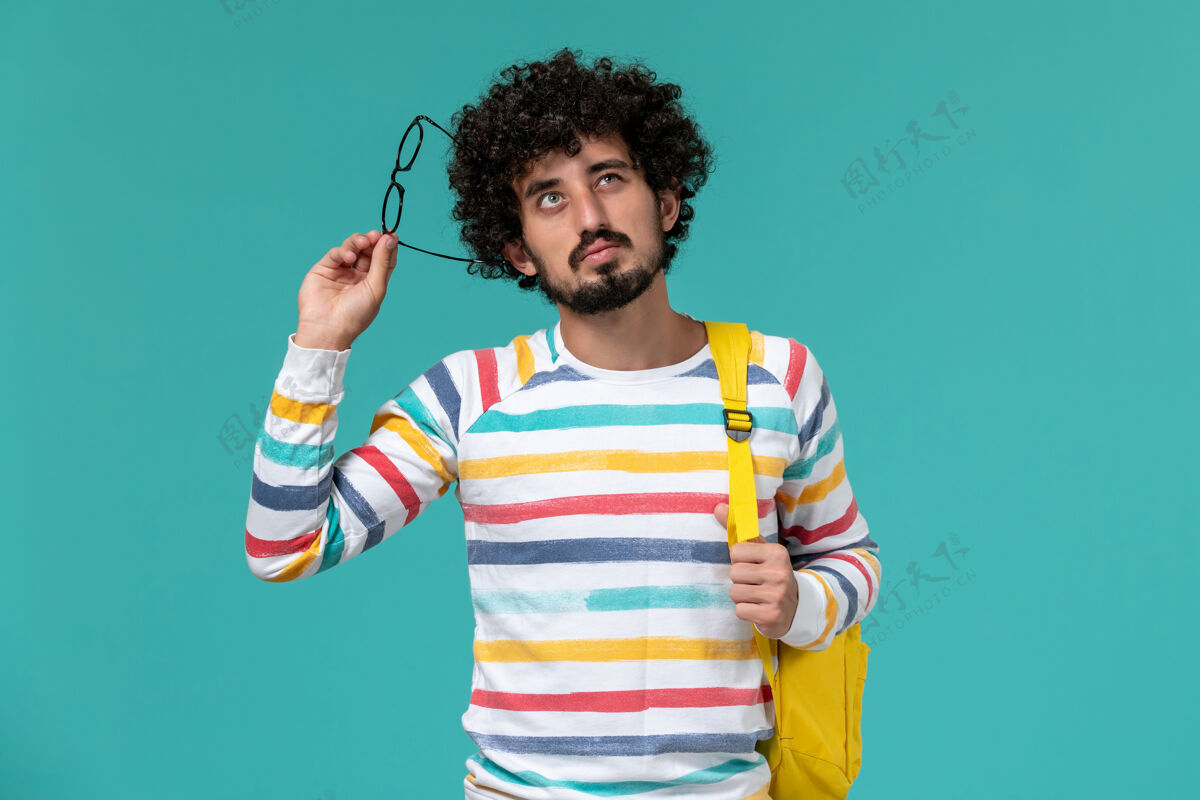 男人穿着条纹衬衫 背着黄色背包 戴着光学太阳镜的男学生在蓝色墙上思考的正视图太阳镜人教训