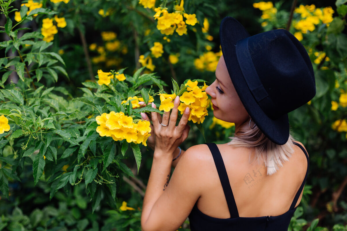 热带美丽时尚的白种人快乐的女人穿着黑色连衣裙 戴着经典的帽子在公园里被黄色的泰国花包围自然可爱灌木